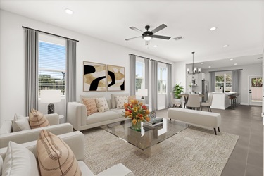 Spacious open‑concept living area with neutral‑colored furniture, large windows with gray curtains, and modern décor. A glass coffee table with flowers sits on a textured rug, and the room flows into a dining area and kitchen with contemporary lighting.